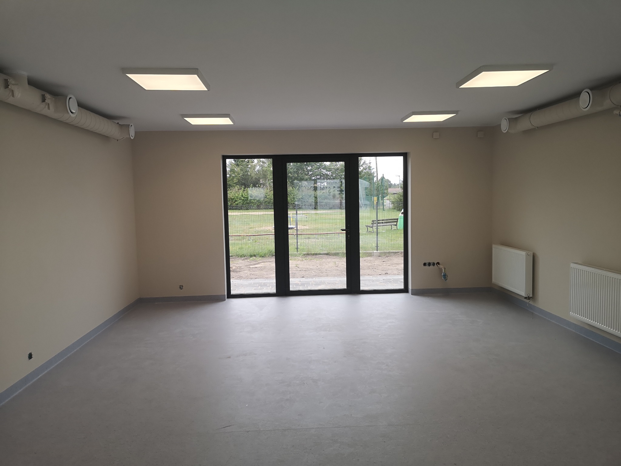 Jasne, puste pomieszczenie z szarym podłożem i dużymi oknami. Widoczne grzejniki, rury wentylacyjne pod sufitem i listwy przypodłogowe. Minimalistyczny wystrój.