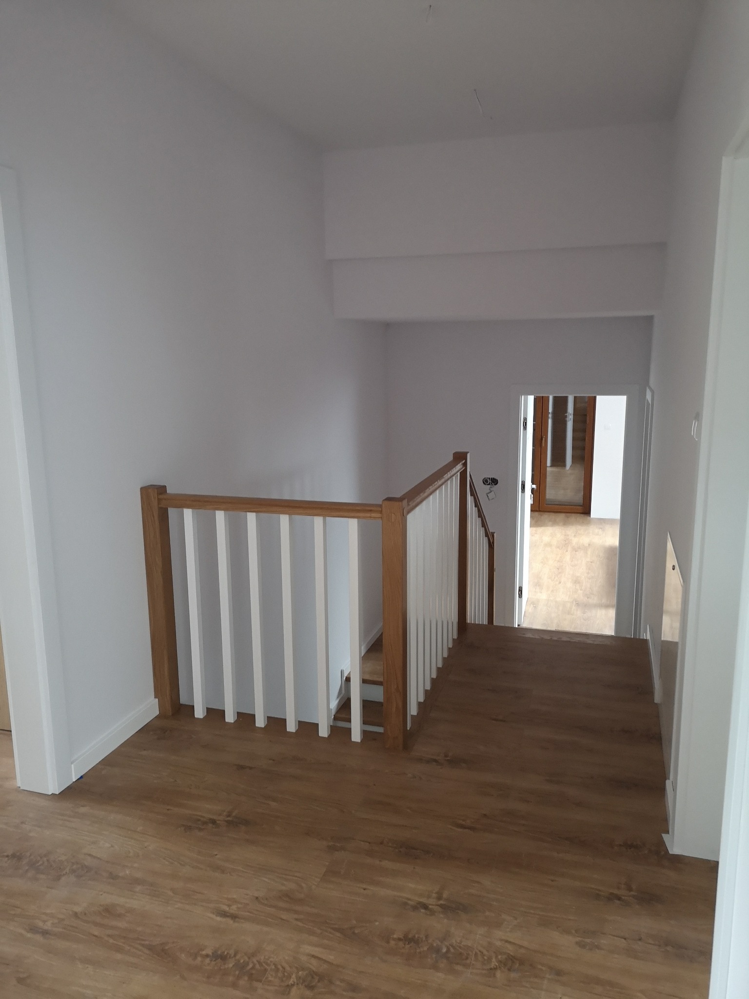 Jasne wnętrze domu po remoncie: białe ściany, drewniane schody z białymi balustradami i panele podłogowe w odcieniach brązu. Widok z góry na dół schodów.