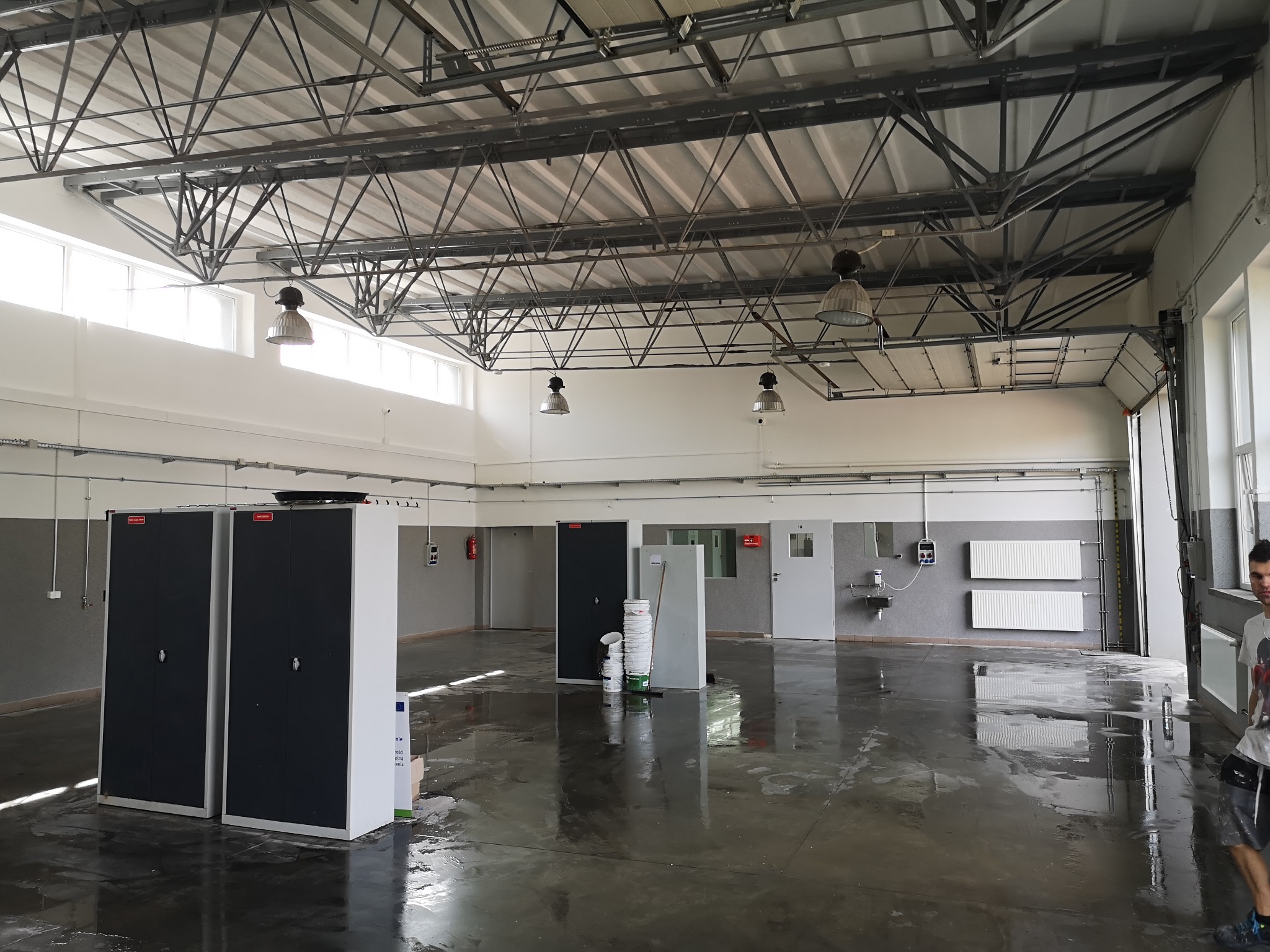 Duże, jasne pomieszczenie po wylewce posadzki. Widoczne szafy, grzejniki, okna i metalowa konstrukcja dachu. Podłoga mokra, odbija światło.