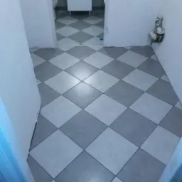 Ułożona podłoga w łazience z płytek w szachownicę, widoczne niedoskonałości przy ścianach, instalacja wodna wystająca ze ściany, w tle umywalka z szafką i toaleta.