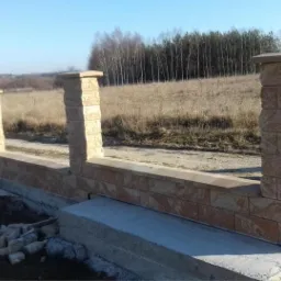 Wznoszona konstrukcja ogrodzenia z kamiennych bloków, z widocznymi słupkami i podmurówką, na tle polnej drogi i lasu w oddali.