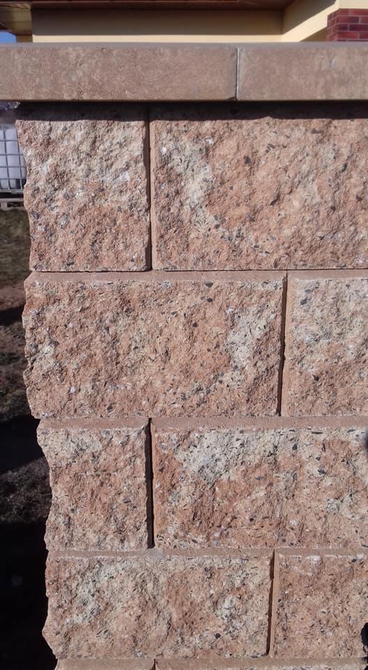 Fragment ogrodzenia z bloków łupanych w odcieniach beżu i różu, z widoczną strukturą i fugami, z lekkim zarysem budynku w tle.