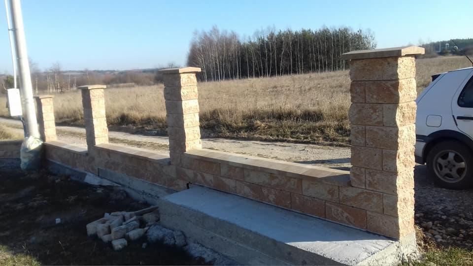 Wznoszona konstrukcja ogrodzenia z kamiennych bloków, z widocznymi słupkami i podmurówką, na tle polnej drogi i lasu w oddali.