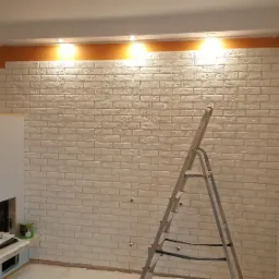 Ściana z białej cegły dekoracyjnej w trakcie montażu, widoczne niedokończone fragmenty, drabina oparta o ścianę, narzędzia na stole roboczym obok kominka.