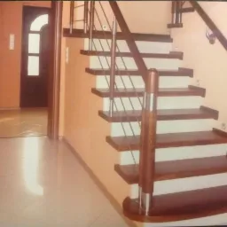 Wewnętrzne schody z drewnianymi stopniami i białymi podstopnicami, balustrada ze stali nierdzewnej i drewnianymi słupkami, widok z dołu, jasna podłoga i drzwi w tle.