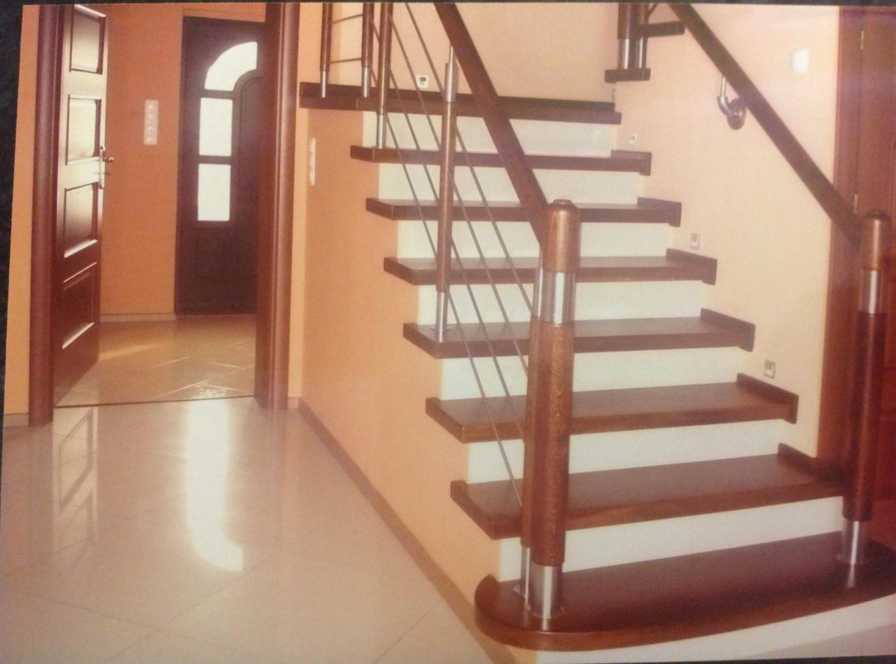 Wewnętrzne schody z drewnianymi stopniami i białymi podstopnicami, balustrada ze stali nierdzewnej i drewnianymi słupkami, widok z dołu, jasna podłoga i drzwi w tle.