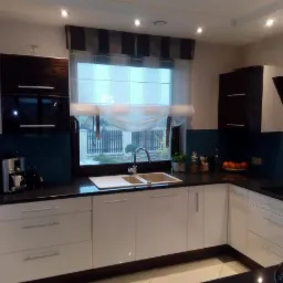 Nowoczesna kuchnia w Nekli z białymi szafkami, ciemnym blatem i niebieskim panelem nad zlewem, widok na ogród przez okno z roletą rzymską.