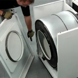 Serwisant w rękawicach pokazuje na otwarte drzwiczki i bęben pralki podczas naprawy.