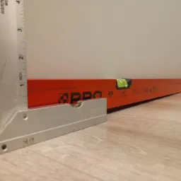 Pomarańczowa poziomica i aluminiowy kątownik na podłodze z paneli, narzędzia do pomiaru i oceny jakości wykonania prac budowlanych.