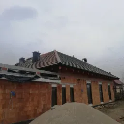 Budowa domu jednorodzinnego z częściowo ułożonym dachem z dachówki ceramicznej. Widoczny mur z cegły dziurawki i sterta piasku na pierwszym planie. Na dachu pracuje dekarz.