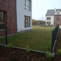 Nowoczesne ogrodzenie panelowe w Szczecinie, oddzielające trawnik od budynku mieszkalnego, z widocznymi słupkami i podmurówką, oraz świeżo posadzone krzewy ozdobne wzdłuż ogrodzenia.