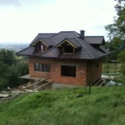 Niedokończony, dwukondygnacyjny dom z czerwonej cegły z ciemnobrązowym dachem, widoczny w pochmurny dzień na wzgórzu, z częściowo wykonaną konstrukcją balkonu i widokiem na oddalone miasto w tle.