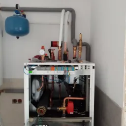 Wnętrze jednostki rekuperacyjnej w trakcie montażu, widoczne rury miedziane, zawory, manometry i podłączenia elektryczne na tle białej ściany, z szarymi rurami instalacji wentylacyjnej u góry.