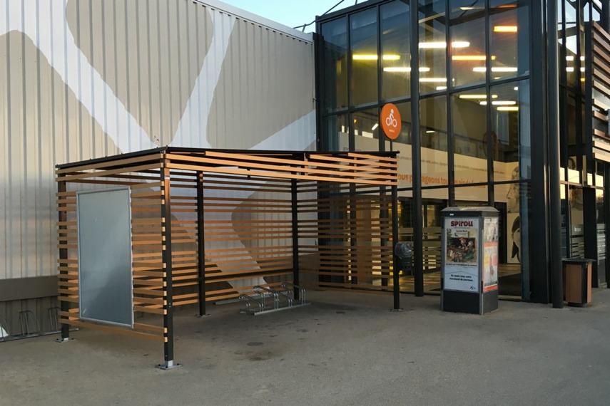 Zadaszona wiata rowerowa z drewnianymi listwami na tle budynku z przeszklonym wejściem.