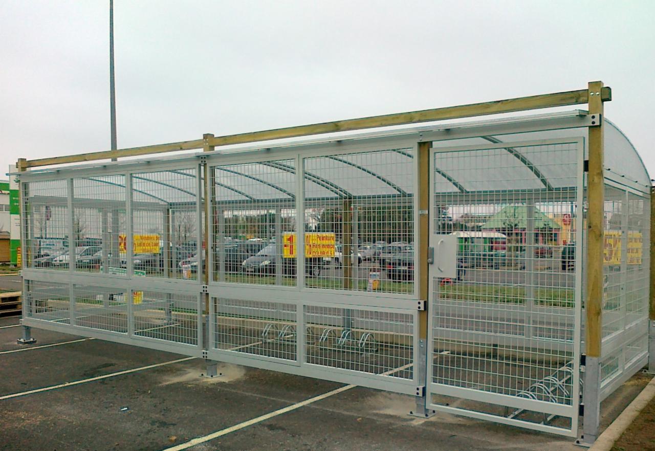 Zadaszona, metalowa konstrukcja z siatki na rowery, z drewnianymi elementami wykończeniowymi, umieszczona na parkingu.