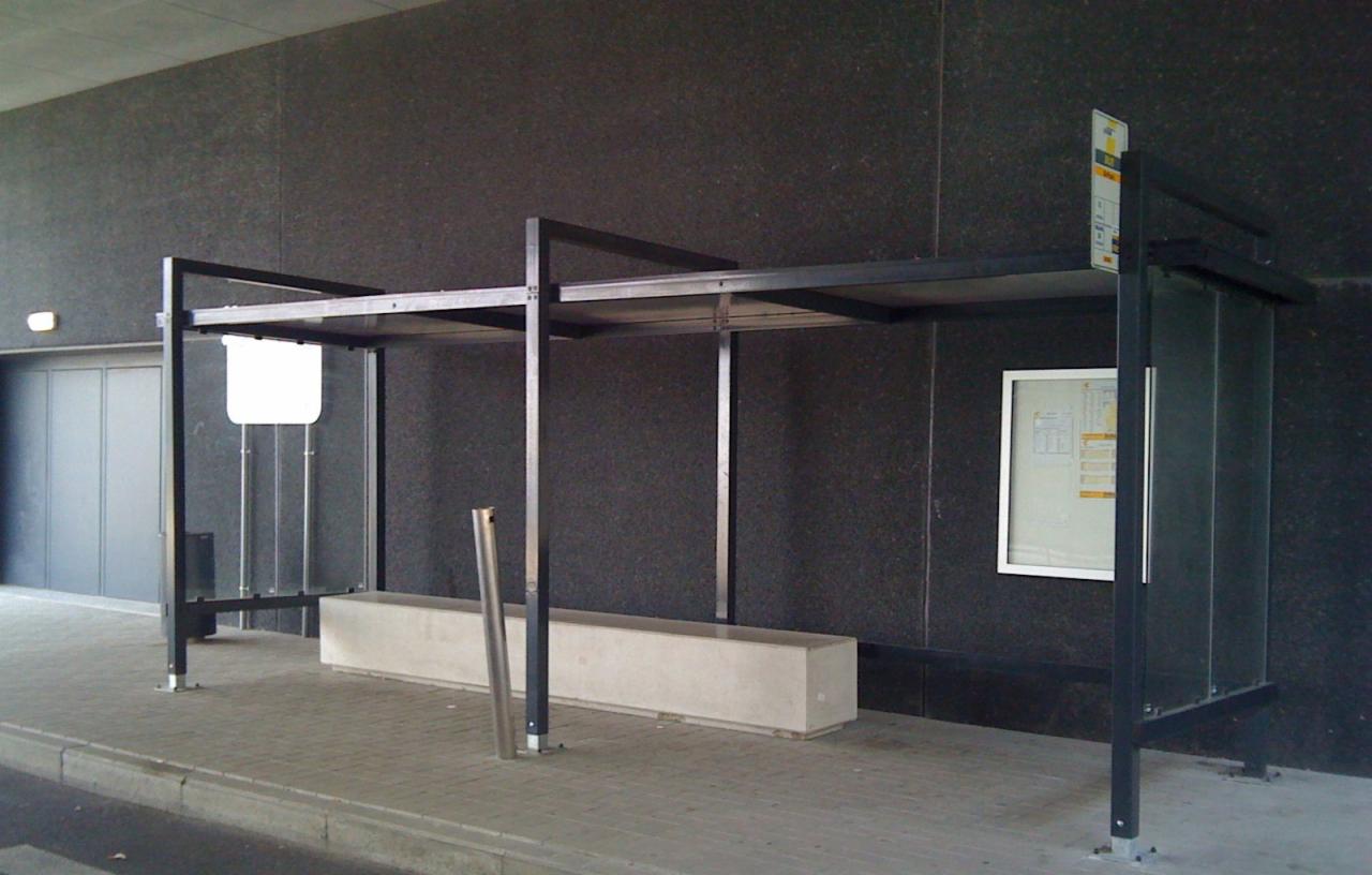 Nowoczesna wiata przystankowa z metalową konstrukcją, betonową ławką i gablotą informacyjną na tle ciemnej, teksturowanej ściany budynku.