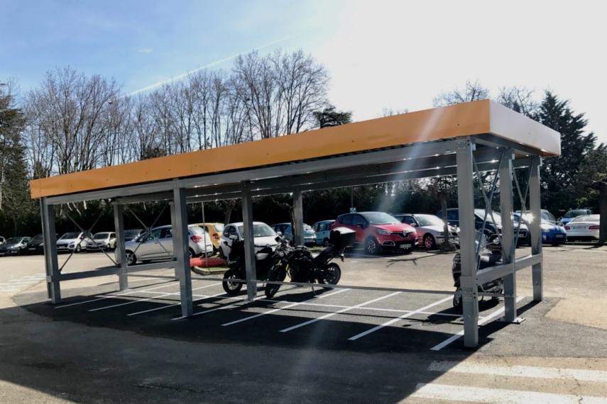 Zadaszone miejsce parkingowe dla motocykli z pomarańczowym dachem na parkingu z samochodami w tle.