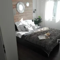 Jasna sypialnia z drewnianą ścianą za łóżkiem, ozdobiona trzema okrągłymi lustrami, na łóżku futrzana narzuta i srebrna taca z filiżankami, podłoga z ciemnego drewna.