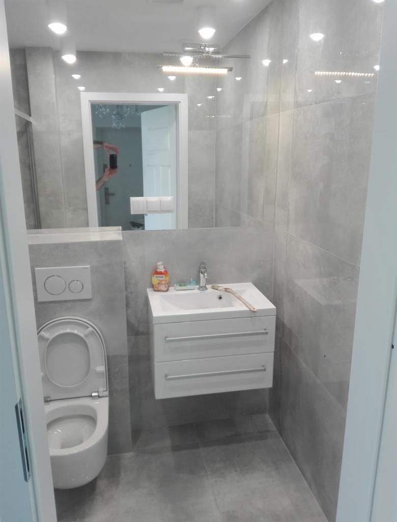 Nowoczesna łazienka w odcieniach szarości z podwieszaną szafką umywalkową, wiszącą toaletą i dużym lustrem z widocznym odbiciem pokoju z żyrandolem.