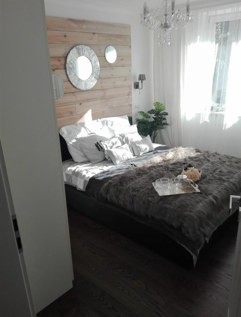 Jasna sypialnia z drewnianą ścianą za łóżkiem, ozdobiona trzema okrągłymi lustrami, na łóżku futrzana narzuta i srebrna taca z filiżankami, podłoga z ciemnego drewna.