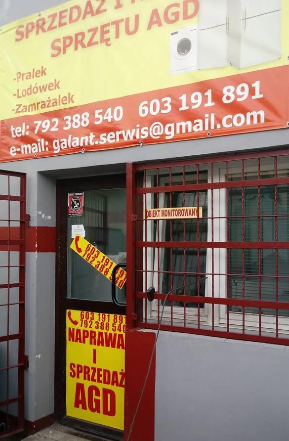 Wejście do punktu naprawy i sprzedaży AGD z reklamą na żółto-czerwonym banerze, obejmującą pralki, lodówki, zamrażarki oraz numery telefonów i adres e-mail, zabezpieczone kratami.