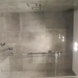 Nowoczesna kabina prysznicowa z szarymi płytkami, minimalistyczną armaturą i szklanymi drzwiami przesuwnymi, widoczna z zewnątrz pomieszczenia.