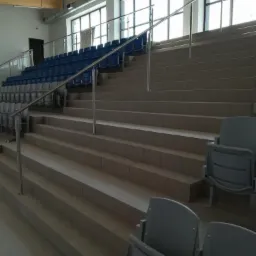 Wygląd schodów i rzędów krzeseł na trybunach hali sportowej, wykończonych płytkami ceramicznymi w odcieniach szarości, z metalowymi barierkami i niebieskimi krzesełkami w tle.