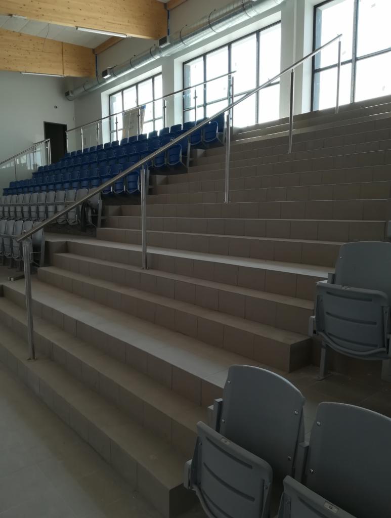 Wygląd schodów i rzędów krzeseł na trybunach hali sportowej, wykończonych płytkami ceramicznymi w odcieniach szarości, z metalowymi barierkami i niebieskimi krzesełkami w tle.