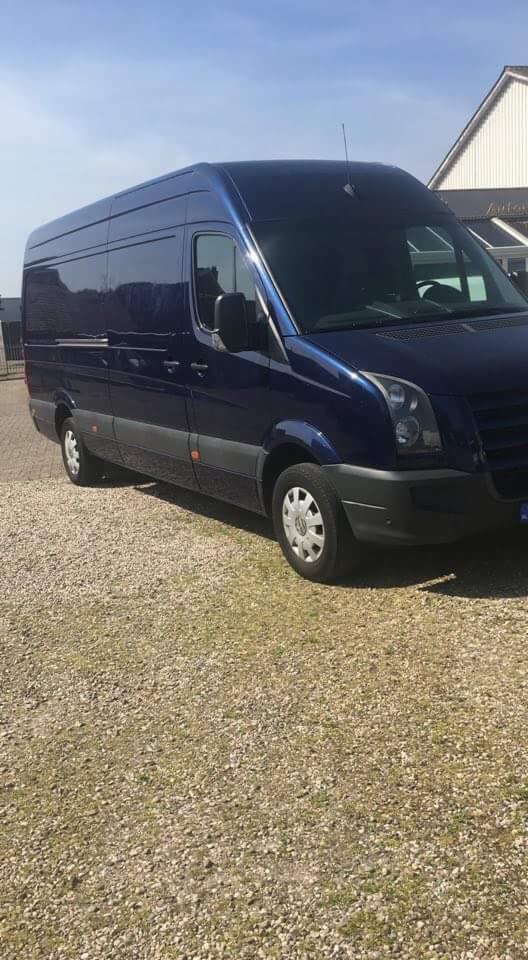 Niebieski, lśniący van dostawczy marki Volkswagen Crafter zaparkowany na żwirowym podłożu w słoneczny dzień. Widoczny budynek w tle z napisem 'Autoteile'.