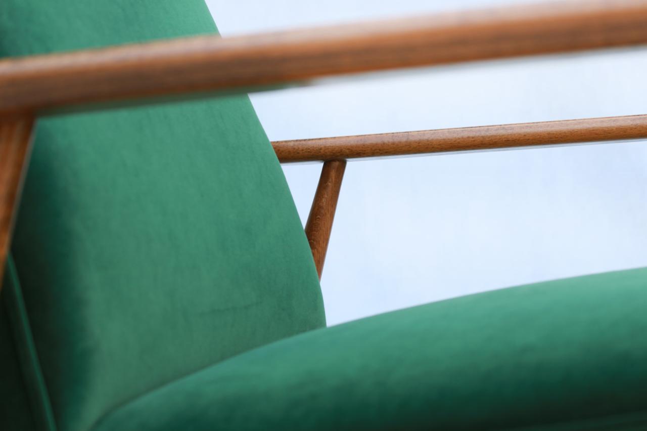 Fragment odnowionego fotela z drewnianymi podłokietnikami i obiciem z zielonego weluru, widoczne delikatne załamania na tkaninie siedziska.