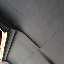 Wooden Houses - Fragment nowego, czarnego pokrycia dachowego z widocznymi łączeniami blach, przy krawędzi okna dachowego, z elementami montażowymi.