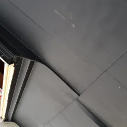 Fragment nowego, czarnego pokrycia dachowego z widocznymi łączeniami blach, przy krawędzi okna dachowego, z elementami montażowymi.