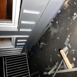 Wooden Houses - Szare obróbki blacharskie okna po montażu, z widocznymi fragmentami blachy na ziemi i niebieskim przewodem wzdłuż ramy okiennej.