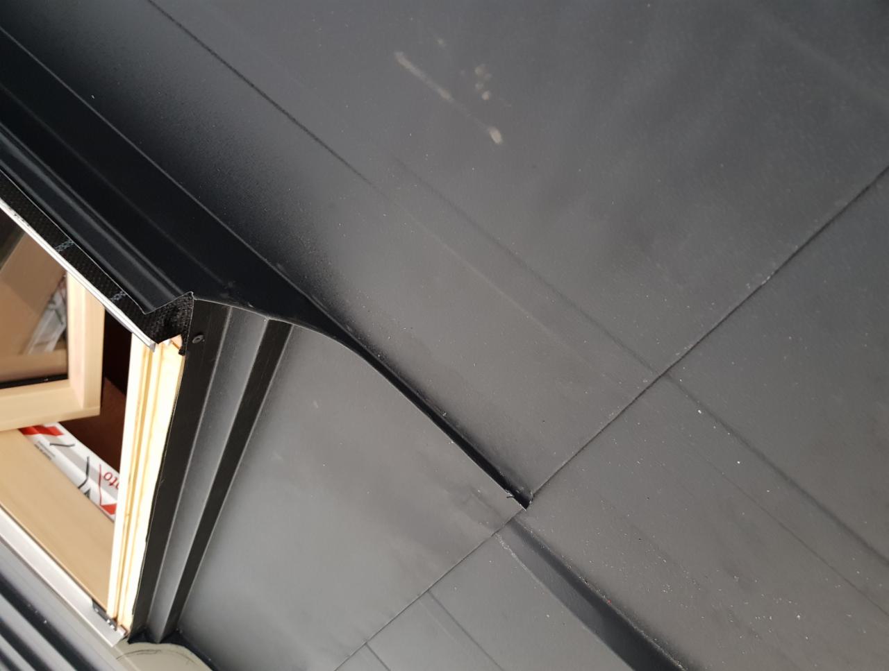 Fragment nowego, czarnego pokrycia dachowego z widocznymi łączeniami blach, przy krawędzi okna dachowego, z elementami montażowymi.