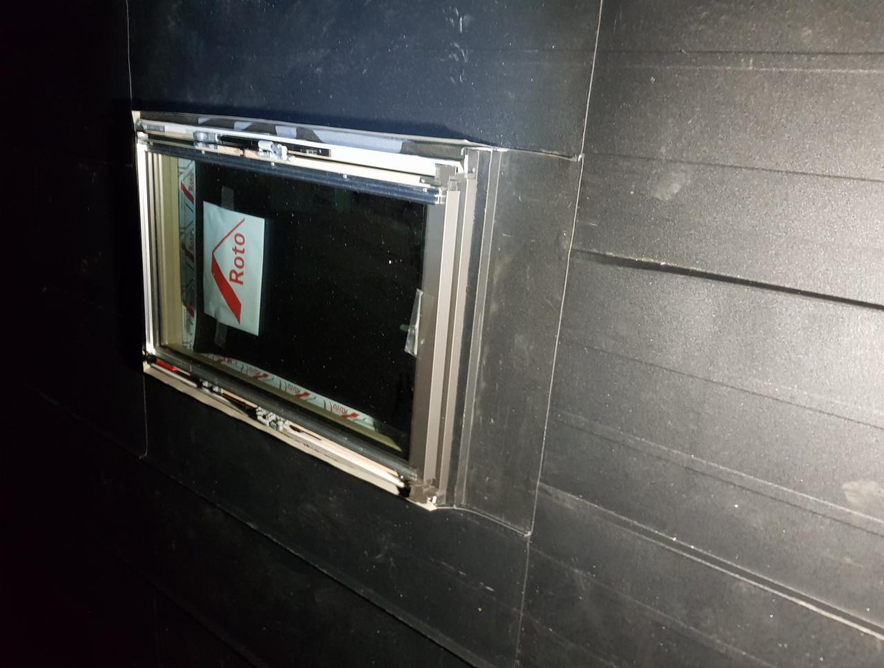 Nowe okno dachowe marki Roto, zamontowane w czarnej, drewnianej konstrukcji dachu, z widoczną folią ochronną na szybie.