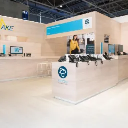 Jasne stoisko targowe z logo i nazwą firmy Sunake i NFON, prezentujące telefony VoIP i monitory, z osobą w tle, oświetlone światłem z góry.