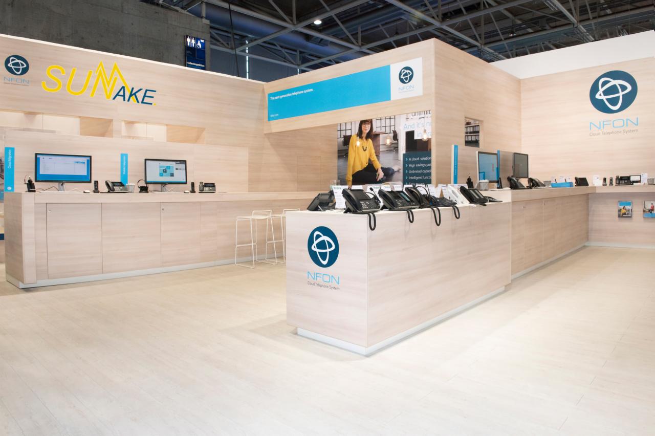 Jasne stoisko targowe z logo i nazwą firmy Sunake i NFON, prezentujące telefony VoIP i monitory, z osobą w tle, oświetlone światłem z góry.