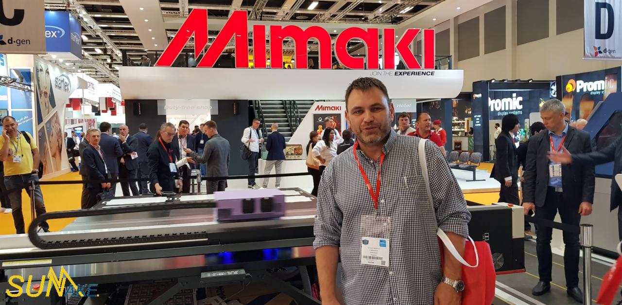 Mężczyzna na tle stoiska firmy Mimaki z ploterem drukującym w trakcie targów branżowych, widoczny ruchomy element z fioletową obudową.