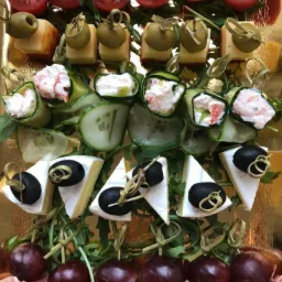 Dekoracyjne koreczki cateringowe na złotej tacy: pomidorki cherry z mozzarellą, ser z oliwką, roladki z ogórka z kremowym nadzieniem, brie z czarną oliwką, winogrona i szynka parmeńska...