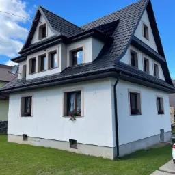 Dwupiętrowy dom z ciemnym dachem i białymi ścianami, małe kwiaty w doniczkach na parapetach, widok z trawnika w słoneczny dzień.