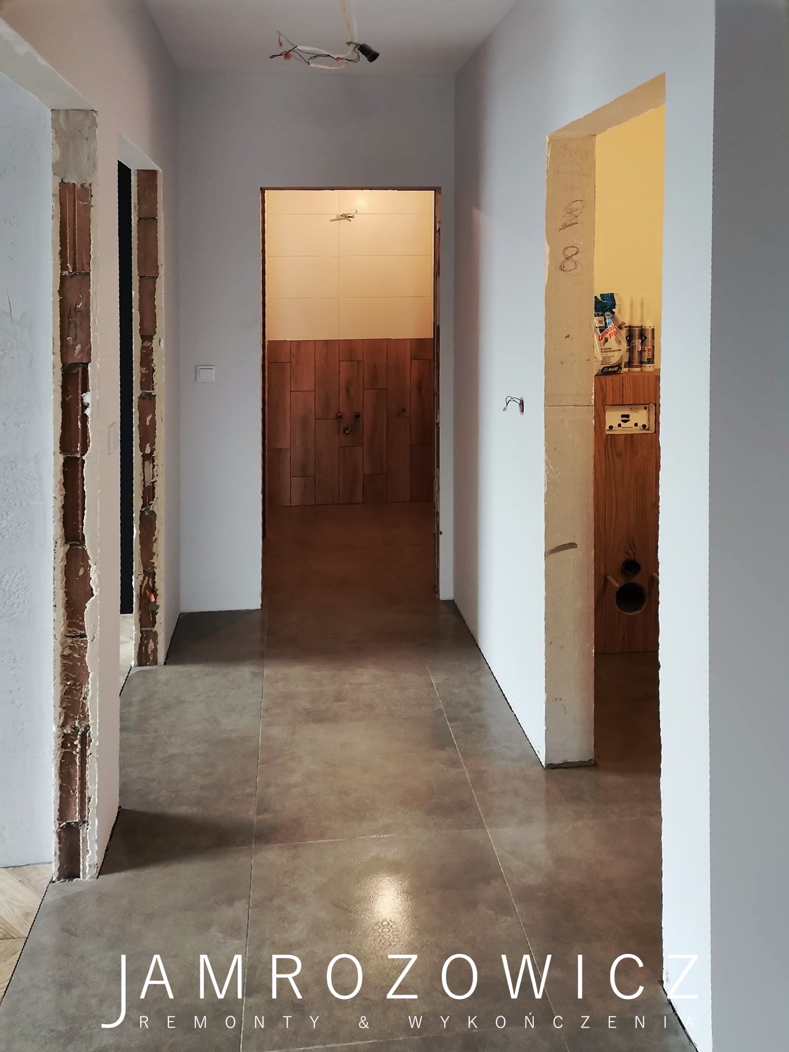 Korytarz w trakcie remontu z widoczną łazienką w tle, świeżo położone szaro-beżowe płytki podłogowe, ościeżnice drzwi przygotowane do montażu, instalacja elektryczna wystająca z sufitu.