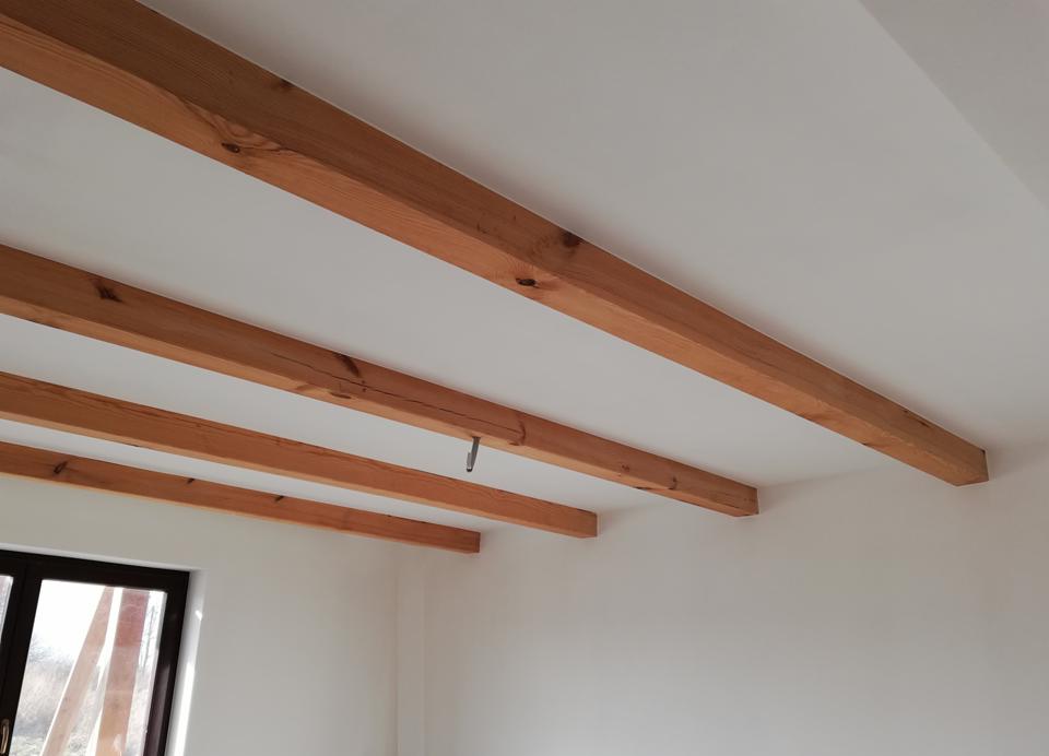 Biały sufit z trzema drewnianymi belkami, widoczne okno w dolnej części kadru, hak zamontowany w jednej z belek.