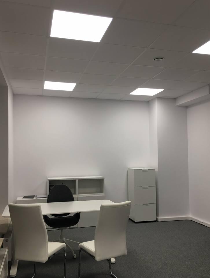 Białe biuro z szarym dywanem, wyposażone w biurko, krzesła i szafki, oświetlone kwadratowymi lampami sufitowymi.