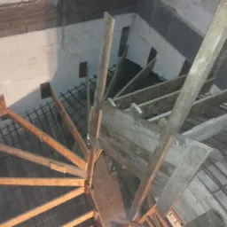 Wewnętrzna konstrukcja spiralnych schodów z drewnianych belek i metalowego zbrojenia, widok z góry, surowy stan budowy w pomieszczeniu z białymi ścianami z betonu komórkowego.