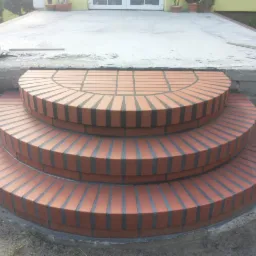 Półokrągłe, trójstopniowe schody wejściowe wyłożone czerwoną cegłą klinkierową z ciemnymi fugami, prowadzące do betonowego podestu przed budynkiem z białymi drzwiami i roślinami w donicach.