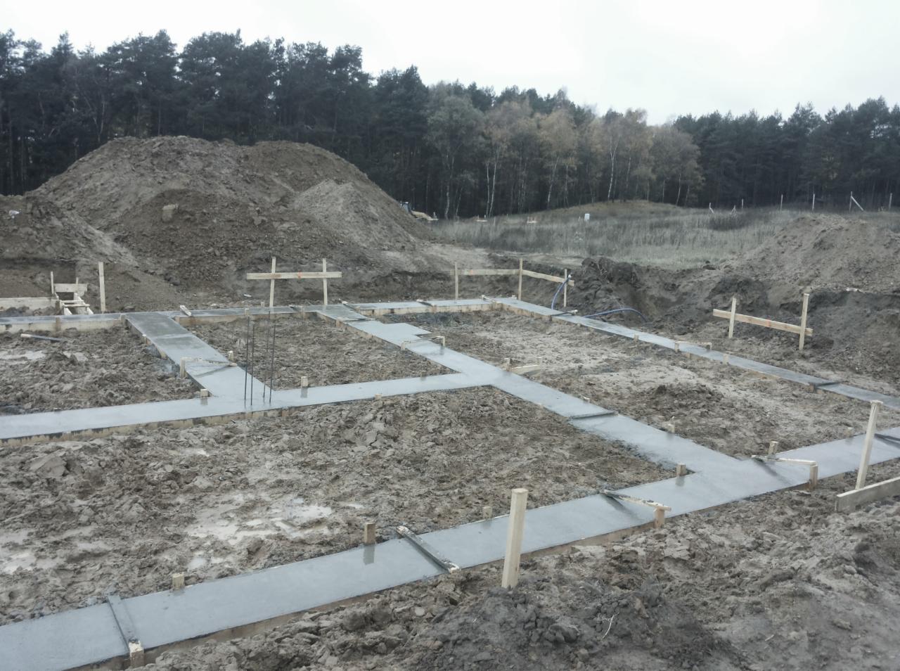 Wylane fundamenty pod budowę domu, widoczne szalunki i zbrojenie, w tle hałda ziemi i las.