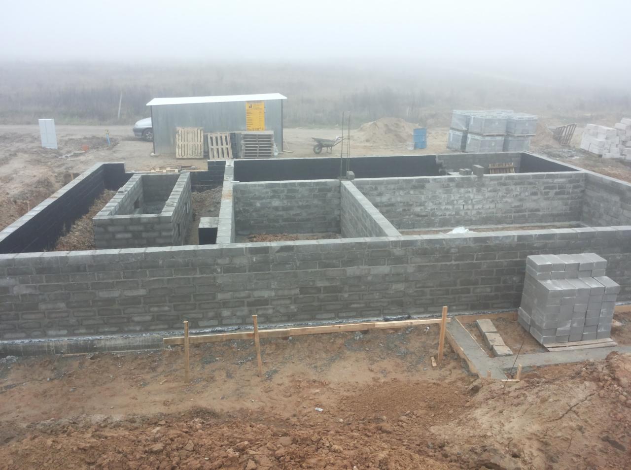 Budowa fundamentów z bloczków betonowych w trakcie realizacji, widoczne zbrojenia i izolacja, w tle zamglony krajobraz i zaplecze budowy z wózkiem i paletami.