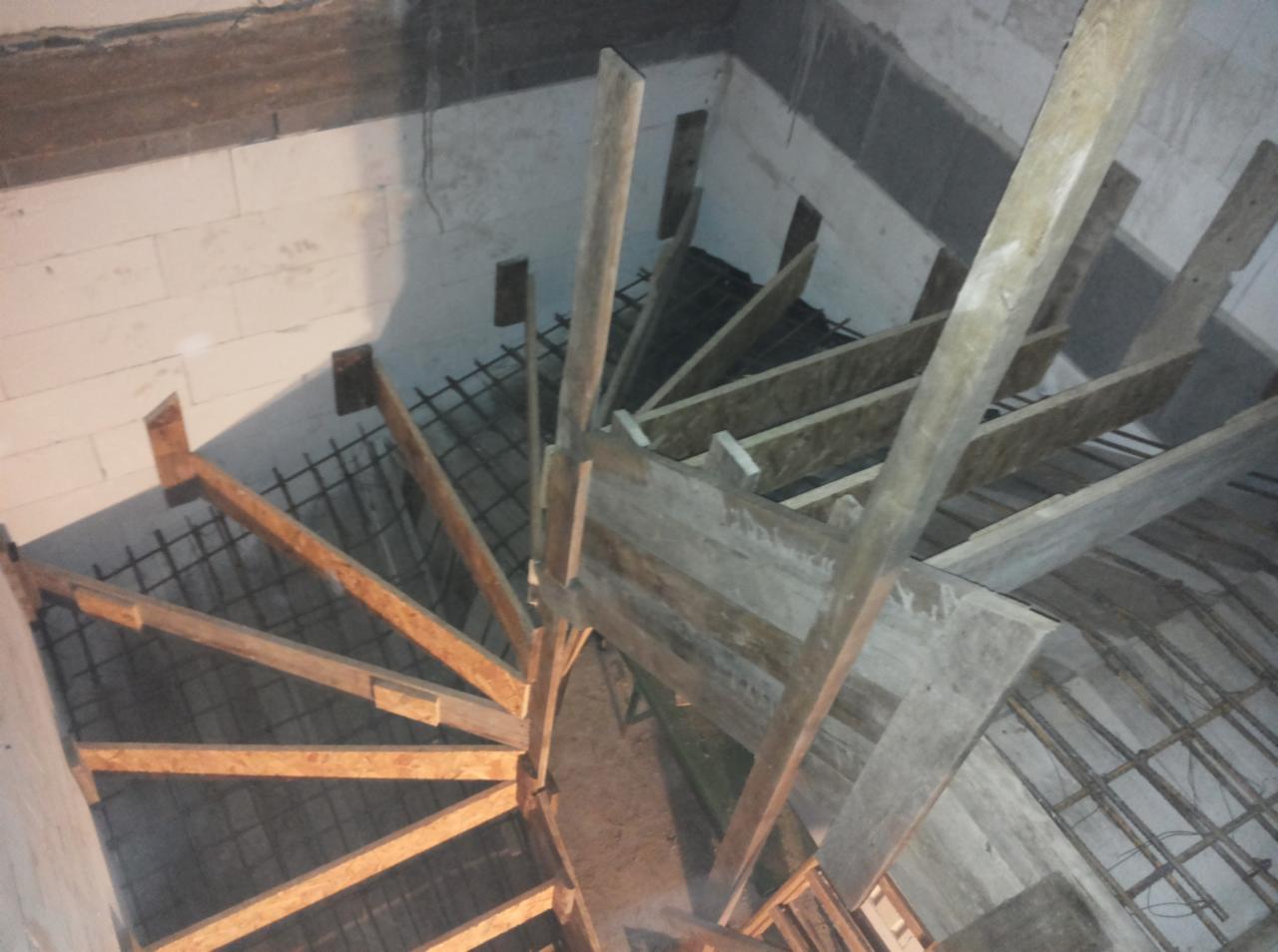 Wewnętrzna konstrukcja spiralnych schodów z drewnianych belek i metalowego zbrojenia, widok z góry, surowy stan budowy w pomieszczeniu z białymi ścianami z betonu komórkowego.