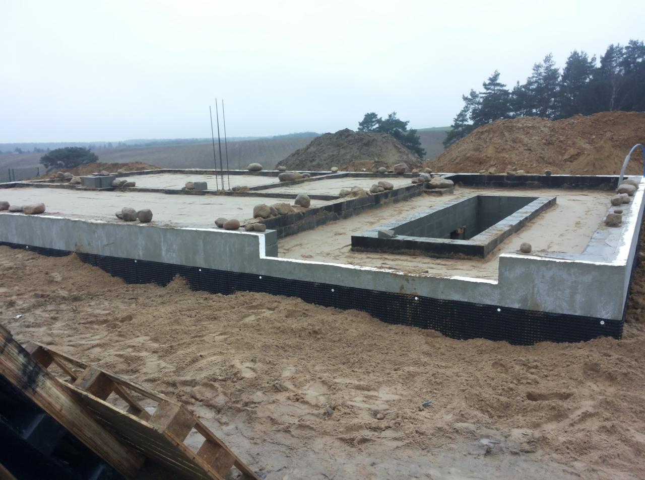 Wylany fundament z izolacją pionową i kamieniami w narożnikach, widoczne pręty zbrojeniowe i wykop pod piwnicę, w tle hałda piasku i drzewa.