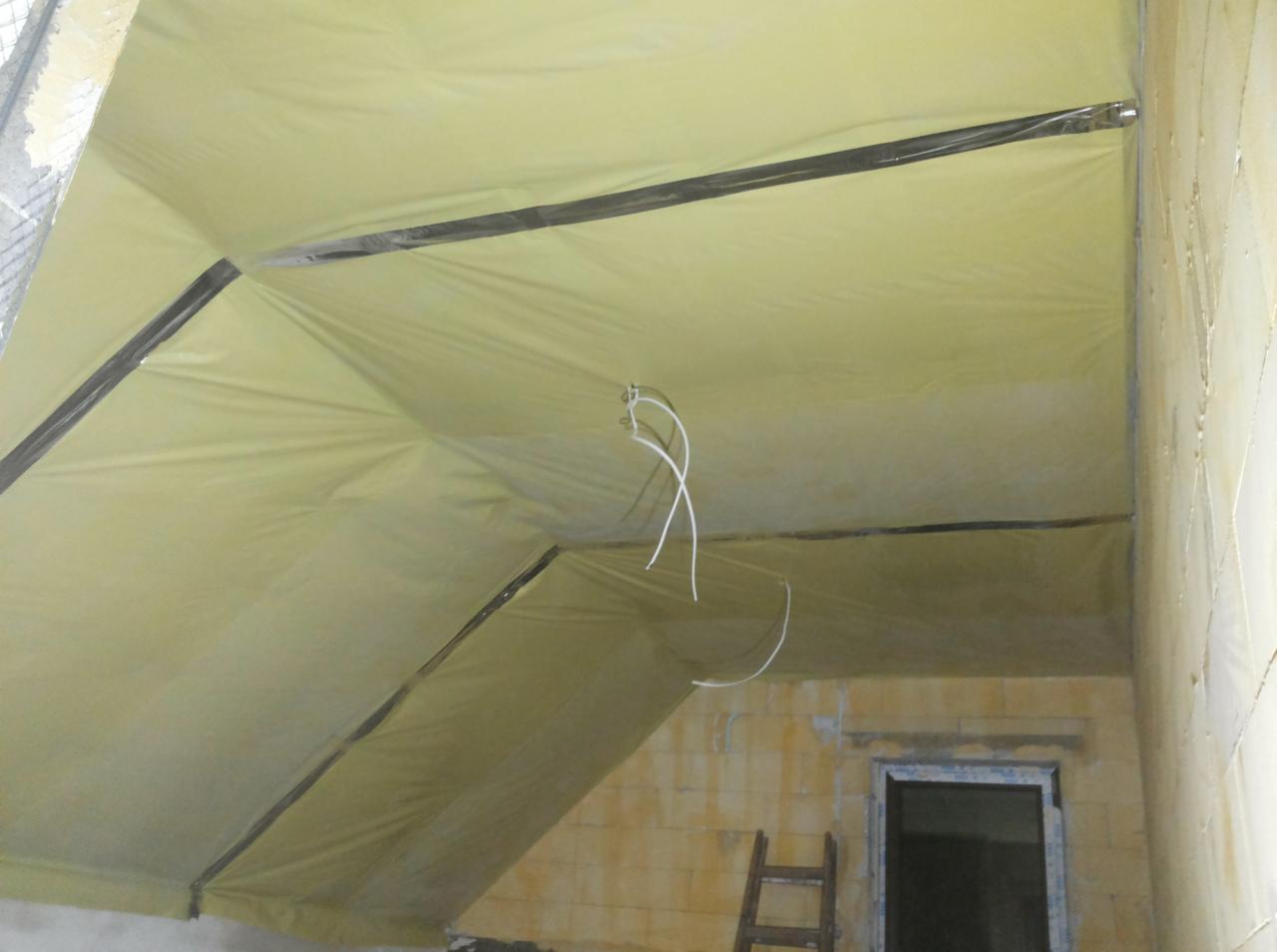 Żółta, rozpięta folia sufitowa z widocznymi profilami montażowymi i wystającymi kablami, w tle surowa ściana z oknem i drabina.